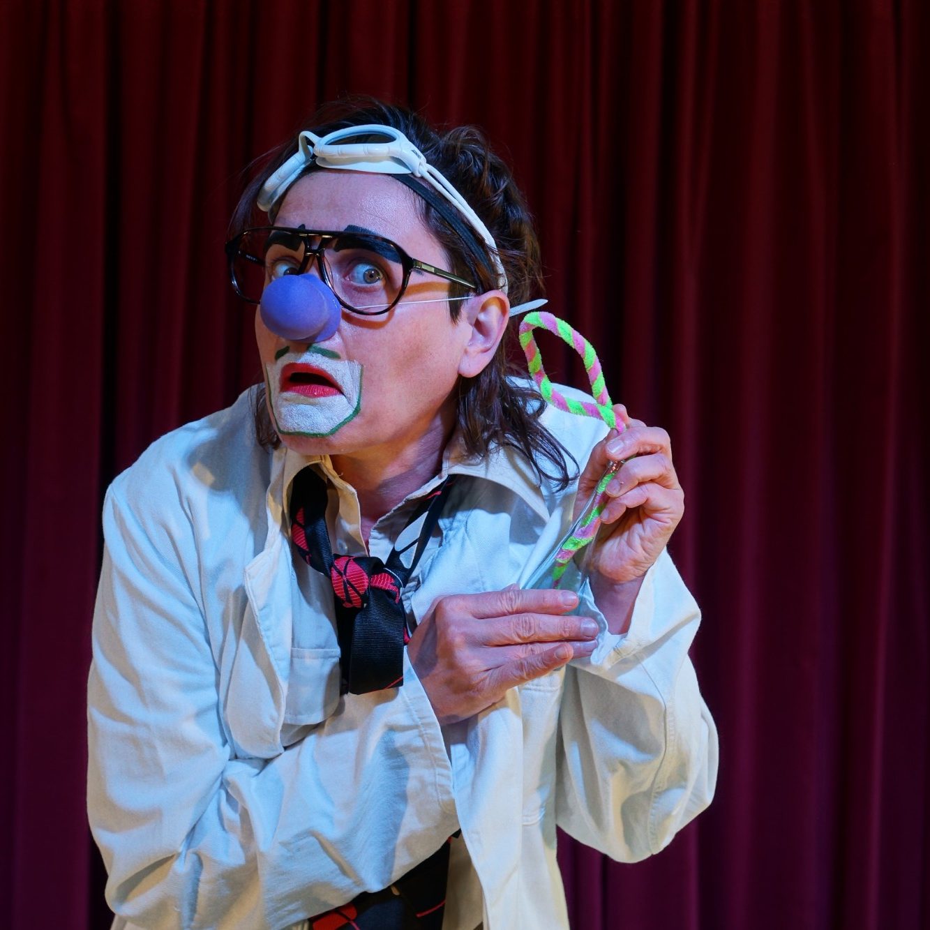 Ein Clown in einem weißen Kittel zeigt eine bunte Süßigkeit mit einem verwirrten Ausdruck.